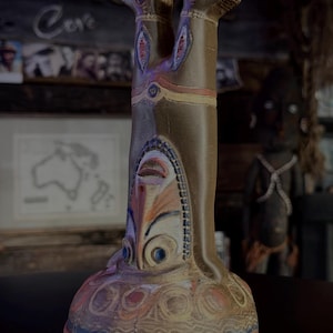 May include: A brown, carved wooden sculpture with a tribal design. The sculpture features a central column with a face-like carving and two upward-reaching arms. The base and arms are decorated with colourful patterns in red, blue, and yellow.