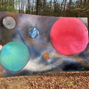 May include: A colorful space-themed painting featuring multiple planets in shades of red, teal, blue, and orange against a dark, starry background. The artwork is signed in the lower right corner. The painting is displayed outdoors.
