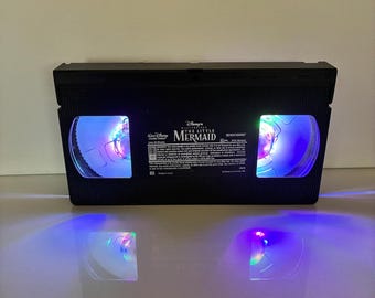 May include: A black VHS video cassette featuring the title "The Little Mermaid" is lit with blue and purple lights. The tape is placed on a reflective surface, creating a mirrored light effect.