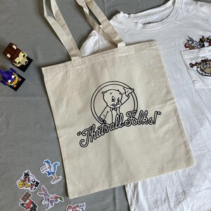 May include: A cream-colored tote bag with the phrase "That's all Folks!" and a cartoon pig illustration. A white t-shirt with a pocket featuring cartoon characters is next to the bag. Several cartoon character stickers are also visible.