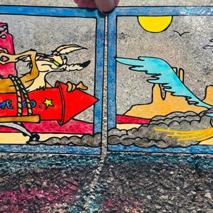 May include: Two-panel artwork featuring cartoon characters. One panel shows a coyote on a red rocket with "ACME CO." text. The other panel depicts a roadrunner running through a desert landscape with a sun and birds.