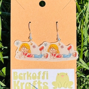 May include: A pair of dangle earrings featuring a cartoon illustration of a girl in a red shirt and blue pants. The earrings are attached to a tan card with the text "Berkoffi Krafts" and a frog graphic.