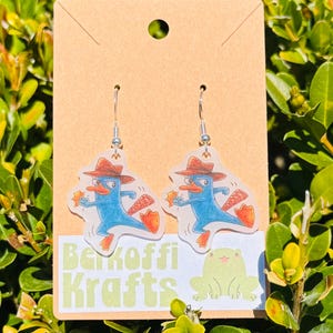 May include: A pair of dangle earrings featuring a cartoon platypus character in a brown hat, holding a star. The earrings are attached to silver-colored fish hook ear wires and are displayed on a brown card with the text "Berkoffi Krafts".