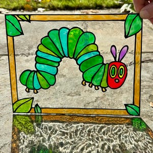 Puede incluir: Un panel de vidriera con un diseño de oruga colorido. La oruga está representada en tonos verdes y turquesas, con una cabeza roja y antenas moradas. El panel está enmarcado con un borde dorado y hojas verdes.