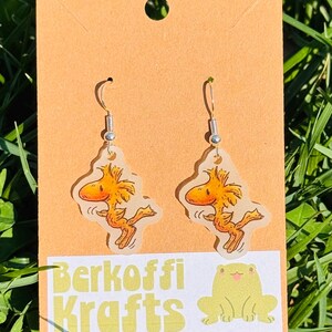 May include: A pair of dangle earrings featuring a cartoon character in shades of yellow and orange. The earrings are displayed on a tan card with the text "Berkoffi Krafts" and a yellow frog graphic. The background is green grass.