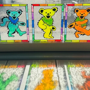 May include: Five colorful stained glass panels featuring dancing bears in blue, green, yellow, orange, and pink. Each bear has a different colored fur and a matching colored collar. The panels are framed with a rainbow border.