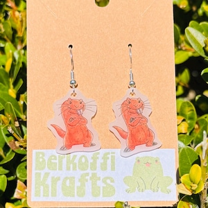 May include: A pair of dangle earrings featuring a cartoon otter design. The otter is orange with a white belly and is drawn with its arms crossed. The earrings are attached to a brown card with the text "Berkoffi Krafts" and a green frog.