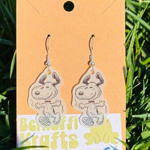 May include: A pair of dangle earrings featuring a cartoon dog character. The earrings are attached to silver-colored hooks and are displayed on a tan card. The earrings have a white background with a cartoon dog design. The text "Belfiffi Krafts" is visible.