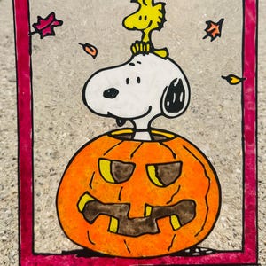 Puede incluir: Panel de vidrio pintado a mano con Snoopy y Woodstock. Snoopy está posado sobre una calabaza naranja tallada, con Woodstock en la cabeza. El panel está enmarcado en un borde rosa, con hojas de otoño dispersas.