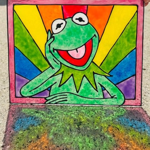 May include: A colorful stained glass panel featuring Kermit the Frog with a rainbow background. The green frog is smiling with his tongue out, framed by a pink border. The panel casts a rainbow shadow.