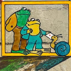 Puede incluir: Vidriera que representa dos ranas antropomórficas y un caracol. Una rana viste ropa marrón, la otra azul. El caracol lleva una carta con un corazón. La obra está enmarcada con un borde amarillo. Ilustración encantadora.