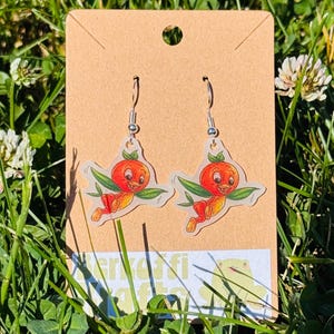 May include: A pair of drop earrings featuring a cartoon orange bird character with green leaves, attached to silver-toned metal hooks. The earrings are displayed on a rectangular tan cardboard backing card with a hole at the top for hanging.