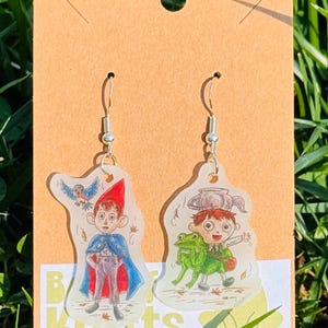 May include: A pair of dangle earrings featuring cartoon characters. One earring depicts a boy in a red hat and blue cape with a bird, the other shows a boy with a frog. The earrings are on a brown card.