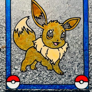 May include: Stained glass art featuring Eevee, a popular Pokémon character. The artwork is framed with a blue border and includes Poké Ball accents in each corner. Eevee is depicted in shades of yellow, brown, and white, with a textured glass background.