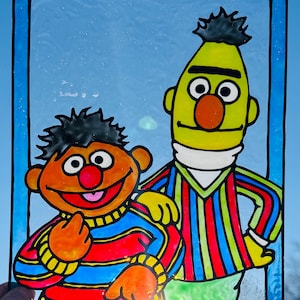 May include: A stained glass panel featuring the characters Bert and Ernie from Sesame Street. Bert is depicted in a green shirt with red, blue, and yellow stripes. Ernie is in an orange shirt with red, blue, and yellow stripes. The panel has a blue border.