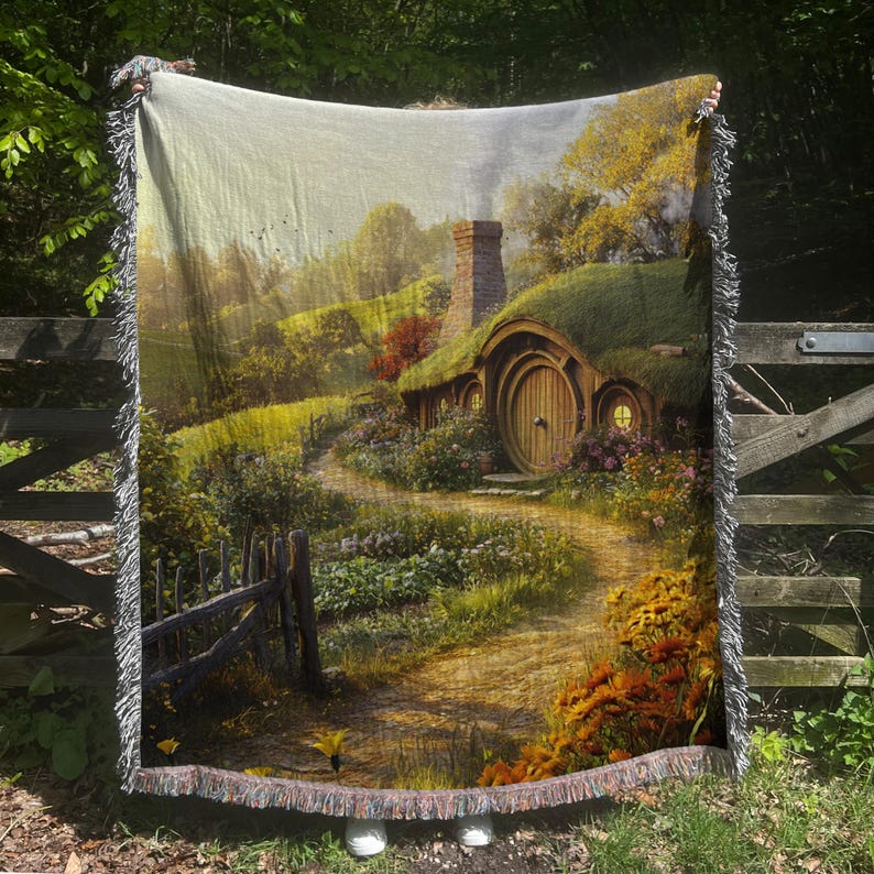 K&ouml;nnte beinhalten: Eine gewebte Wandteppich-Decke mit einer fantasievollen Illustration eines Hobbit-Hauses mit einer runden T&uuml;r, umgeben von &uuml;ppigem Gr&uuml;n und einem gewundenen Weg. Das Haus hat ein Strohdach und einen gemauerten Schornstein.
