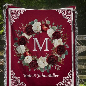 Peut inclure: Plaid bordeaux avec un motif de couronne florale. La couronne présente des roses blanches et bordeaux autour d'une grande lettre blanche "M". La couverture a des franges blanches et les noms "Kate & John Miller" sont imprimés en bas.