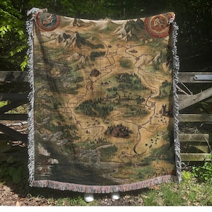 May include: A woven tapestry throw blanket featuring a detailed map of a fictional land. The map is in shades of brown, green, and beige, with a winding road and various landmarks. The blanket has a fringed edge.