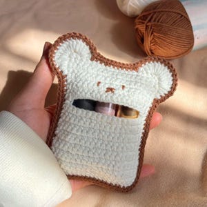 May include: A crocheted pouch in the shape of a slice of bread, with a white centre and brown edging. The pouch has a bear face design. The background shows balls of yarn.