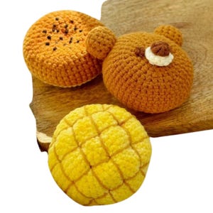 Pode incluir: Três peluches de crochê em forma de pão. Um é um pão marrom em forma de urso, outro é um pão amarelo com um padrão de grade e o último é um pão marrom com sementes pretas no topo.