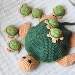 Peut inclure: Une tortue en crochet faite à la main, avec une carapace verte et un corps beige, entourée de petites tortues. La tortue a des yeux en boutons noirs et est posée sur une surface blanche. Le crochet est détaillé et les couleurs sont douces.