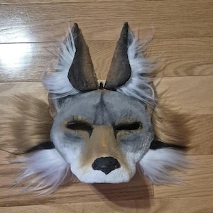 May include: A handmade wolf mask with gray, brown, and white fur accents. The mask features pointed ears, a detailed snout, and eye openings. The mask is made of a textured material and is designed to be worn.