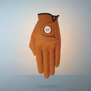 May include: A tan golf glove with a black trim and a circular patch featuring the initials "AC". The glove has small perforations for breathability and is made of a smooth leather material. The glove is designed for right-handed use.