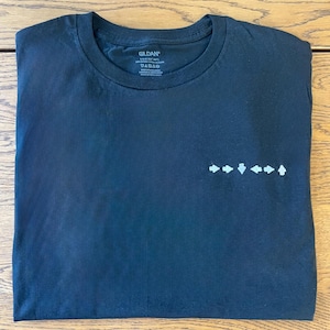 May include: A folded black t-shirt with a crew neck. The shirt has a small, white arrow design on the front. The Gildan brand label is visible at the neckline. The shirt is made of a soft, comfortable material.