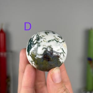 May include: A polished moss agate sphere featuring a white base with intricate, dark green and earthy brown dendritic inclusions that resemble mossy patterns. The letter D is printed in purple above the stone sphere.