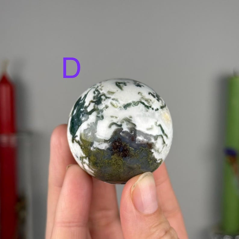 May include: A polished moss agate sphere featuring a white base with intricate, dark green and earthy brown dendritic inclusions that resemble mossy patterns. The letter D is printed in purple above the stone sphere.