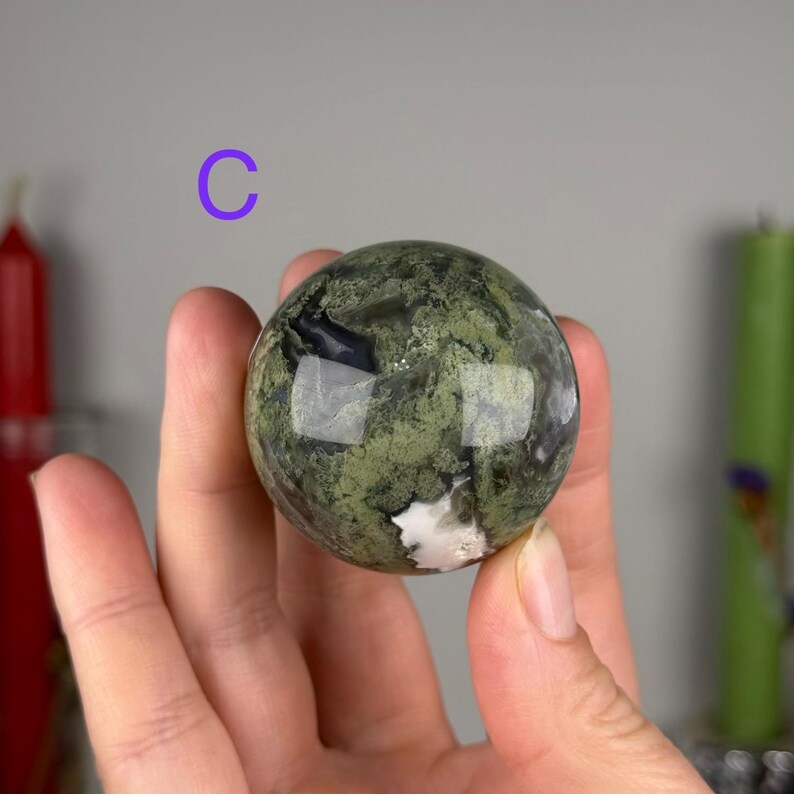 May include: A polished spherical stone featuring a mottled pattern of olive green, dark forest green, and black mineral inclusions with a small patch of white. The sphere is marked with the letter C in purple text positioned above the object.