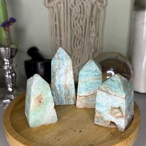 May include: Four polished gemstone towers in shades of blue, green, and brown are displayed on a small wooden tray. The crystals have unique patterns and textures, with a natural, earthy aesthetic. The tray is on a small wooden table.