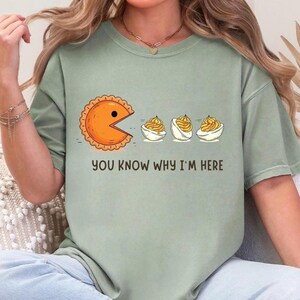 May include: Sage green t-shirt featuring a Pac-Man-inspired pie character chasing three deviled eggs. The text "YOU KNOW WHY I'M HERE" is printed below the graphic. The shirt is a comfortable, casual style.