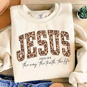 May include: Cream-coloured sweatshirt featuring the word "JESUS" in large, leopard-print letters. Below, the text reads "John 14:6 the way the truth the life" in a script font. The sweatshirt has a crew neck.