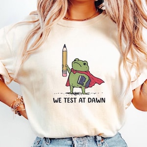 Puede incluir: Sudadera de color crema con un dibujo animado de una rana superhéroe sosteniendo un lápiz y un libro. La rana lleva una capa roja y el texto "WE TEST AT DAWN" está impreso debajo.