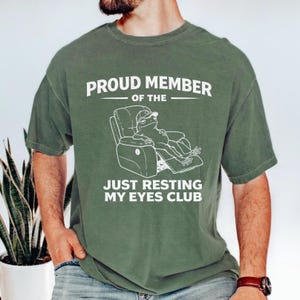 Op de afbeelding: Een olijfgroen t-shirt met korte mouwen en een witte afbeelding van een kikker met een honkbalpet die in een relaxfauteuil zit. De tekst PROUD MEMBER OF THE JUST RESTING MY EYES CLUB staat in witte hoofdletters boven en onder de afbeelding.