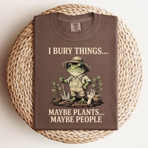 May include: A brown t-shirt with the text "I Bury Things... Maybe Plants... Maybe People" and a cartoon frog wearing overalls and a hat, holding a shovel. The shirt is folded on a woven mat.