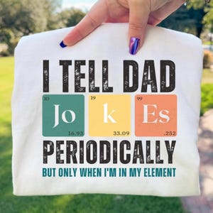 以下が含まれることがあります： 白いTシャツに黒い文字で「I TELL DAD JOKES PERIODICALLY BUT ONLY WHEN I'M IN MY ELEMENT」とプリントされています。JOKESという単語は、原子番号と重量が記載された、青緑色、黄色、オレンジ色の3つの周期表の元素記号風のデザインになっています。