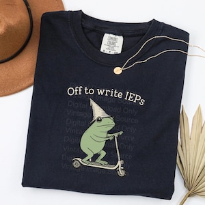 May include: Navy blue t-shirt with the text "Off to write IEPs" and a cartoon frog wearing a wizard hat riding a scooter. A gold necklace and a brown hat are also in the image.