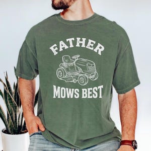 Può includere: T-shirt verde con la scritta bianca "FATHER MOWS BEST" sopra e sotto una grafica bianca di un tosaerba. La maglietta ha maniche corte e girocollo.