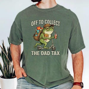 May include: A sage green t-shirt featuring a graphic of a cartoon frog wearing a brown cap, riding a scooter, and carrying a backpack filled with snacks. The frog holds a drink and fries. The text reads OFF TO COLLECT THE DAD TAX in beige.