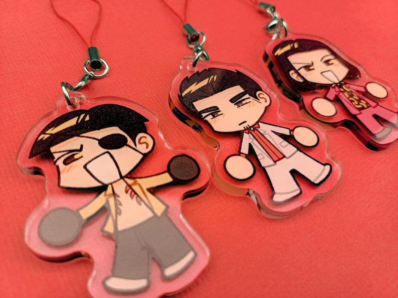 May include: Three acrylic charms featuring anime-style characters with various outfits and expressions. Each charm is attached to a red string with a metal clasp. The characters have black hair and are set against a pink background.