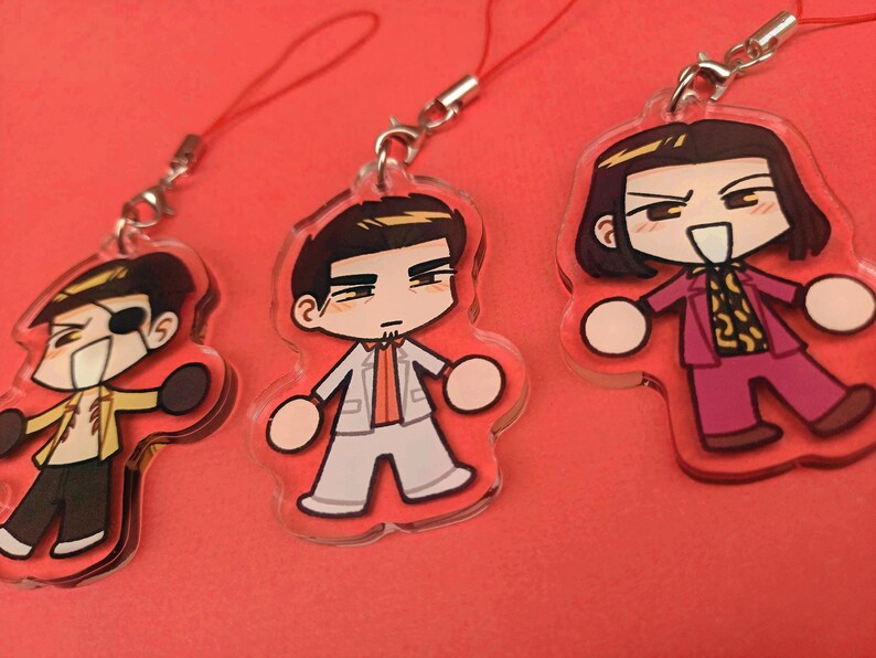 May include: Three acrylic charms featuring cartoon characters. One character wears a yellow jacket and has an eyepatch. Another is in a white suit, and the third is in a pink suit with a patterned shirt. Each charm has a red lanyard.