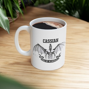 May include: White ceramic mug with a black handle, filled with dark coffee. The mug features a graphic of a muscular figure with wings, the name "Cassian" above, and "Lord of Bloodshed" below.