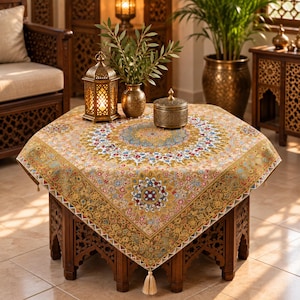 Könnte beinhalten: Eine quadratische Tischdecke mit einem komplizierten, mehrfarbigen Blumen- und Geometriemuster in Gold-, Blau- und Rottönen. Der Stoff zeigt ein zentrales Medaillon-Design, eine dekorative Bordüre und eine kleine Quaste an jeder Ecke.