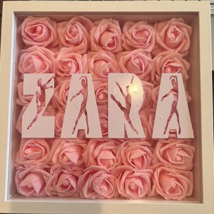 May include: A white framed shadow box filled with pink roses. The word "ZARA" is displayed in the center, with each letter featuring a ballerina silhouette. The roses are densely packed, creating a textured background.