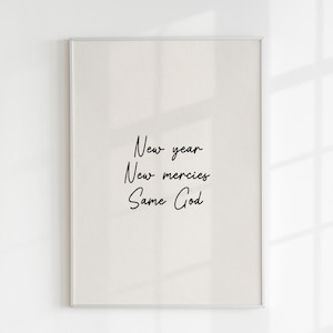 May include: Framed print with the words "New year, New mercies, Same God" in black script on a cream background. The print is in a silver frame and is displayed against a white wall with shadows.