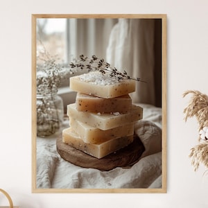 Può includere: Una pila di cinque saponi quadrati di colore chiaro con scaglie visibili, sormontati da rametti e cristalli di sale. I saponi poggiano su una piccola tavola di legno rotonda, su uno sfondo morbido dai toni neutri. L'immagine è incorniciata in legno chiaro.