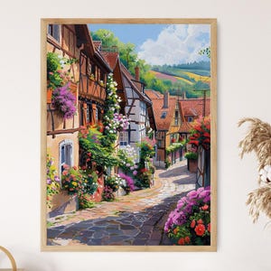 May include: A framed print of a European village street scene. The buildings are half-timbered with red tile roofs and adorned with colorful flowers. The cobblestone street leads into the distance under a blue sky with fluffy clouds.
