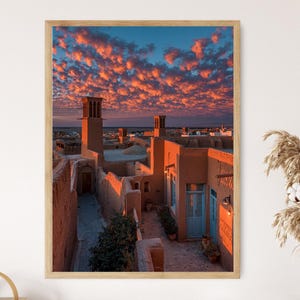 May include: A framed print of an ancient Middle Eastern city at sunset. The image features buildings with wind towers, a blue sky, and vibrant orange and pink clouds. The architecture is a warm, earthy tone.